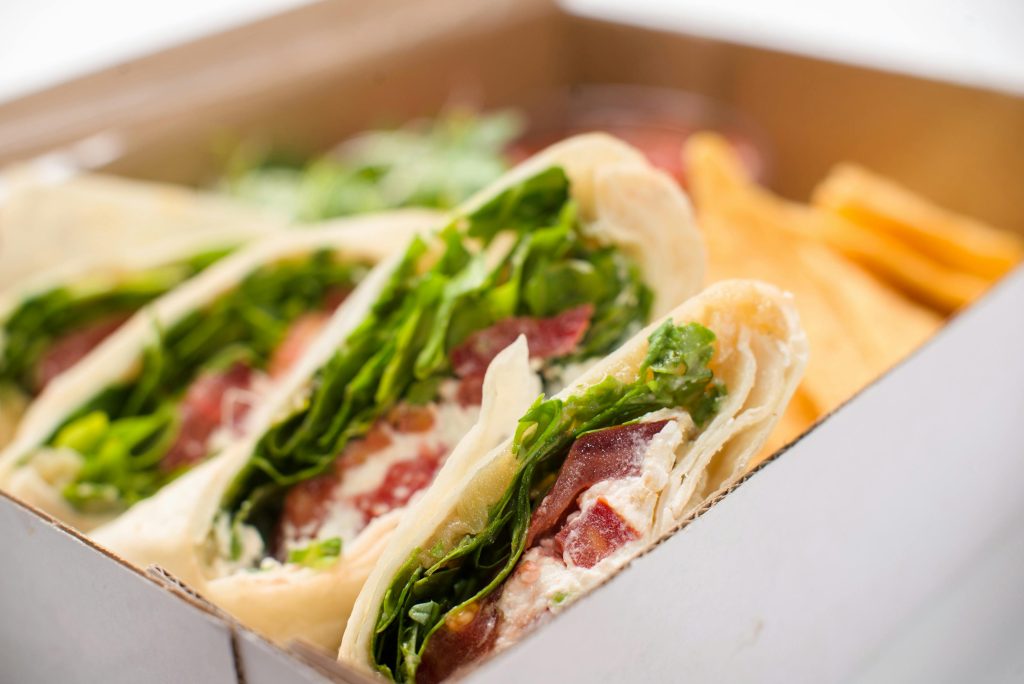 Delicious wrap sandwich featuring greens, bacon, and cheese in a take-out box.